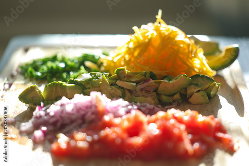 Fresh chopped and grated taco ingredients including tomato, onion, avocado, jalapeno pepper, and cheddar cheese on a cutting board ready to be put in a shell and eaten for dinner. 