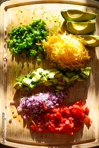 Fresh chopped and grated taco ingredients including tomato, onion, avocado, jalapeno pepper, and cheddar cheese on a cutting board ready to be put in a shell and eaten for dinner. 