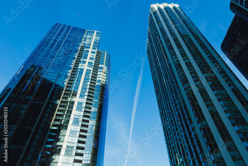 View of tall buildings reaching the sky with a clear blue background and a trail in the air showing a plane flying high above the city