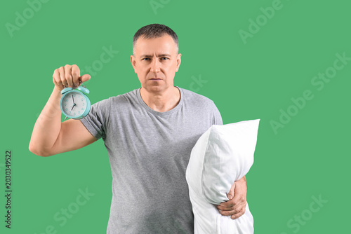 Handsome mature angry man in stylish pajamas with pillow and alarm clock on g...