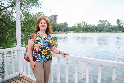 Schilderij op canvas A senior hispanic woman smiles and looks at the camera as she enjoys her free time in a park by a lake