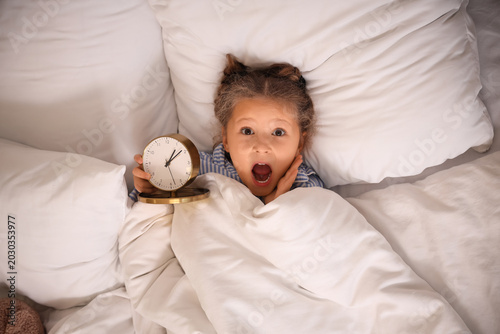 Photos Shocked little girl with alarm clock lying in bed, top view