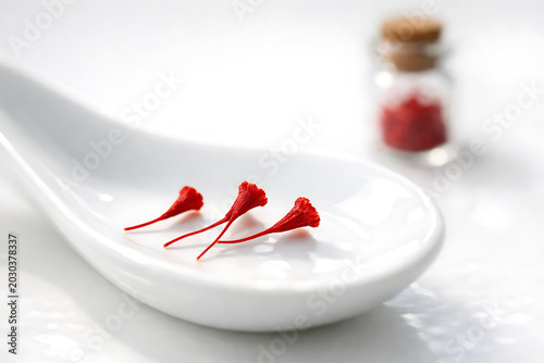 Three delicate crimson saffron threads on a white ceramic spoon with a glass vial