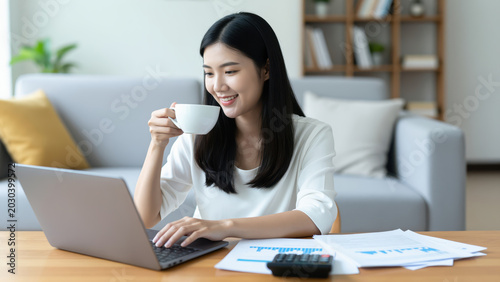Home Office Comfort: A young woman enjoys a coffee break while working on her laptop at a desk, surrounded by documents and a calculator, in a cozy home setting.