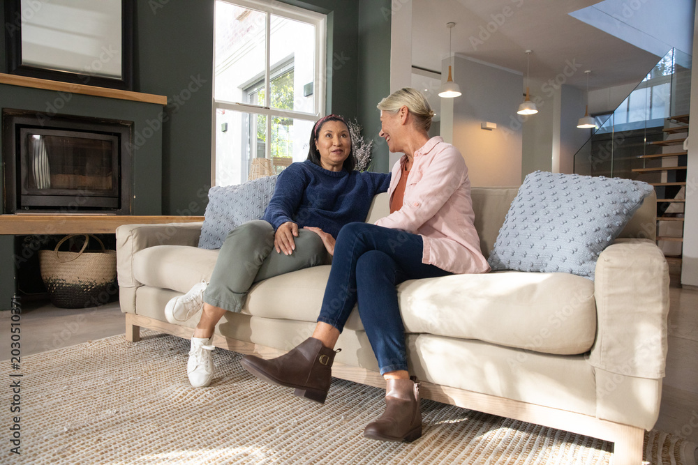 Fototapeta premium Two female partners sitting on beige sofa at home, leaning, smiling, wearing boots and sneakers