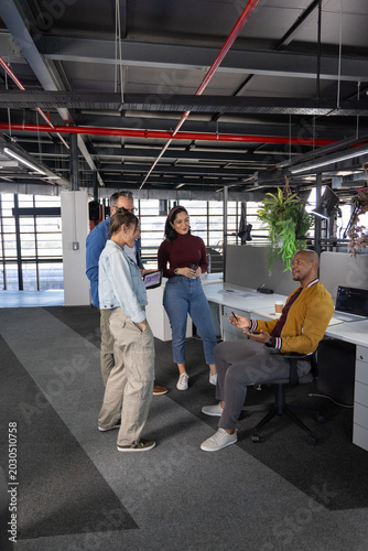 Diverse coworkers discussing project while gathering at desks in modern office with laptop, tablet
