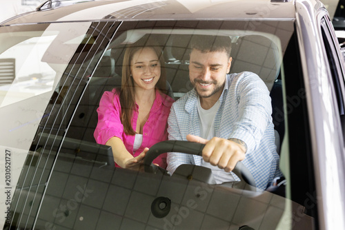 Young couple testing vehicle interior and controls together while sitting inside modern automobile at dealership showroom, viewed through windshield