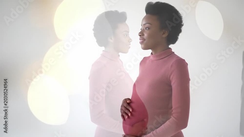 Young pregnant woman in white t-shirt gently cradles bump, serene smile, gazing hopefully in a pristine white studio with bokeh background. Concept of motherhood and anticipation