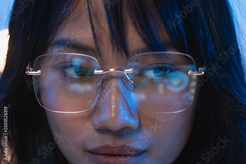 Macro close up portrait of a young Asian female programmer wearing modern rimless eyeglasses with bright digital code reflection visible on the transparent lenses