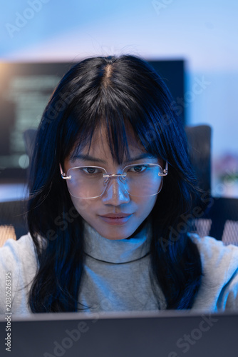 Vertical portrait of a focused young Asian female software developer wearing rimless glasses working on her laptop with a dark monitor and bright code visible behind her