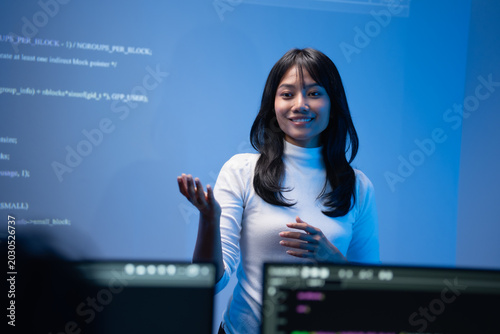 Happy young Asian female programming instructor in a white turtleneck smiling and gesturing with her hand while teaching a coding class in front of a blue digital code wall