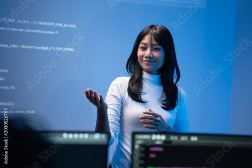Cheerful young Asian woman tech instructor in a white turtleneck sweater smiling brightly while gesturing and teaching programming concepts in front of a blue code wall