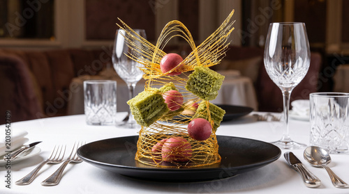 Luxury fine dining dessert with intricate spun sugar art, matcha green tea cubes, and raspberry chocolate truffles on a black plate in a fine restaurant setting
