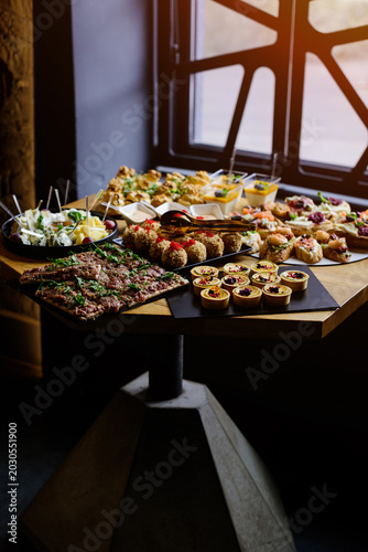A wide shot of a luxurious catering buffet table featuring gourmet appetizers, including meat tartare, salmon and shrimp bruschettas, cheese platter, and mini desserts lit by warm natural light.