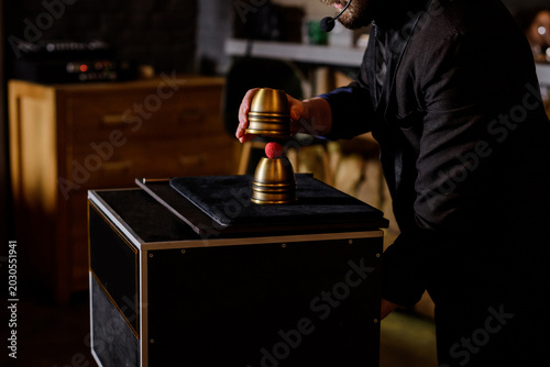 A professional magician performing a classic cups and balls magic trick with golden metal cups and a red ball during a live entertainment show at an event.