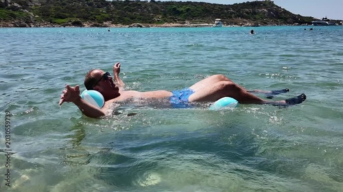 Close-up of a mid-adult man wearing sunglasses, relaxing and sunbathing on a striped inflatable mattress while floating on clear sea water waves.