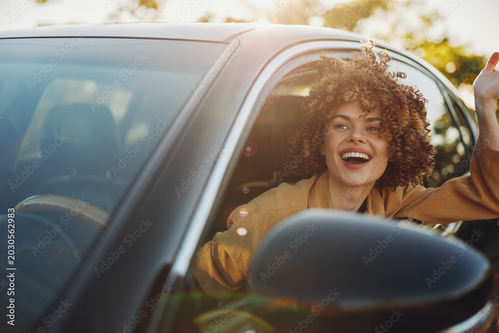 Fototapeta premium A joyful woman in a car with window down, hair in the breeze, bathed in warm retro tones. A candid lifestyle moment that conveys joy, travel, and sunny freedom. Perfect for lifestyle and travel