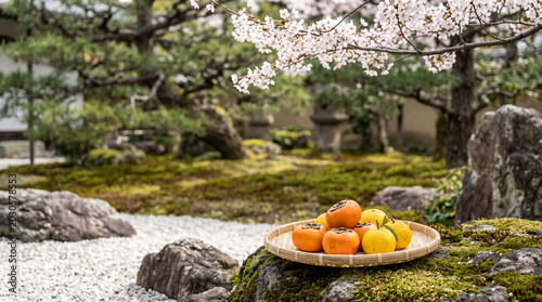 Traditional Japanese Garden Still Life with Fresh Persimmons and Citrus Fruits in Bamboo Basket on Mossy Stone with Cherry Blossoms and Zen Landscape Background