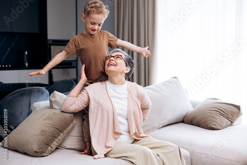 фотографія Grandson standing on couch behind his happy grandmother, both smiling during playtime at home