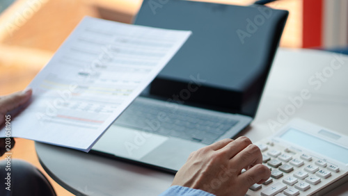 Financial accounting budget and business tax auditing concept. Close up of a person's hands holding a printed report document and typing on a calculator next to a laptop.