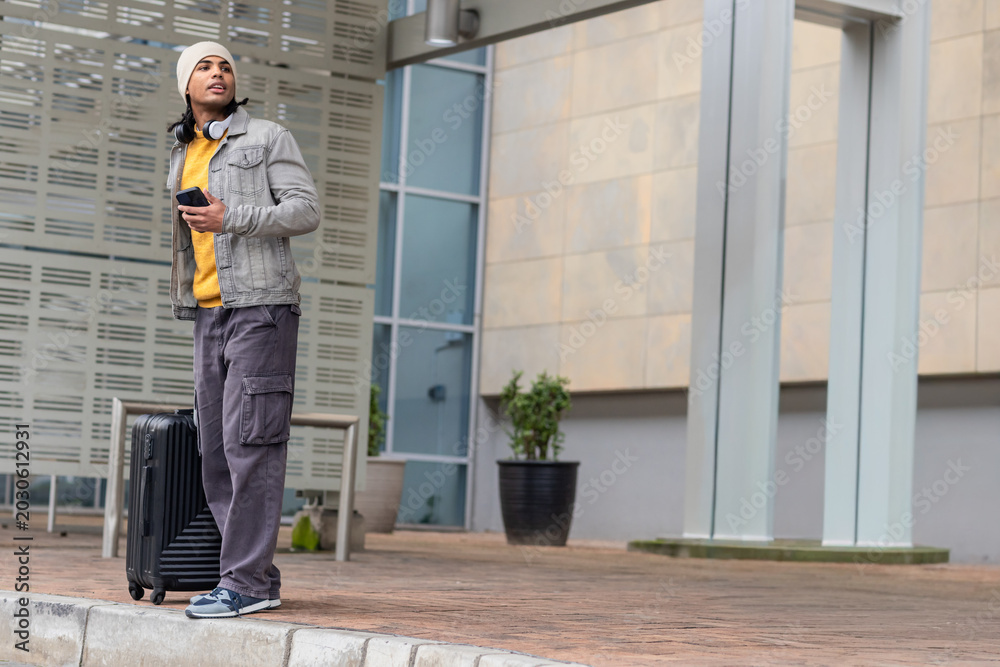 Fototapeta premium Man in his 20s standing on curb outside modern building holding smartphone and suitcase, copy space
