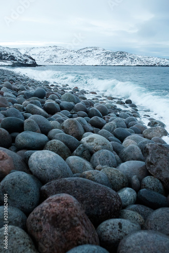 Vertical shot of rocky beach with round stones in winter Teriberka Arctic ocean coast background with snow covered hills and sea waves, Barents sea shore, northern cold dramatic nature travel concept