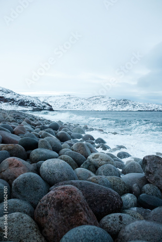 Vertical shot of rocky beach with round stones in winter Teriberka Arctic ocean coast background with snow covered hills and sea waves, Barents sea shore, northern cold dramatic nature travel concept