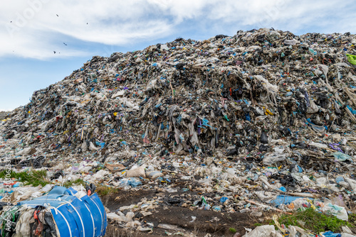 Large mounds of waste and plastic debris are seen at a landfill site close to a road, with birds flying overhead during the day
