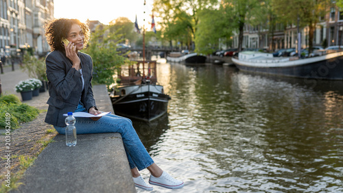 Woman working remotely, talking on phone in amsterdam, enjoying flexible lifestyle by canal at sunset