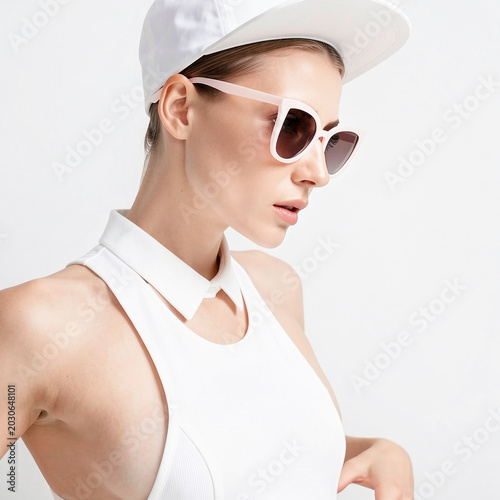 Side profile of trendy young woman in white baseball cap, sunglasses and sleeveless collar shirt against white background.
