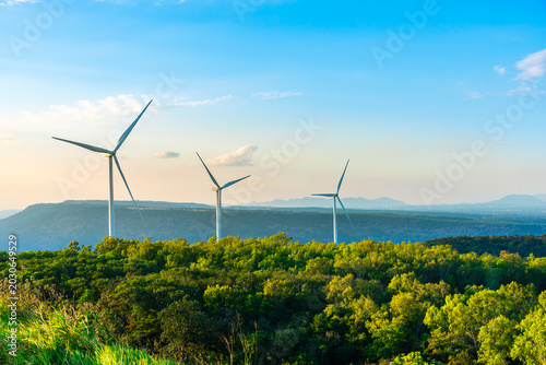 Three large white wind turbines generating clean renewable energy on a lush green hill in Thailand. Industrial eco-friendly power plant landscape technology concept under a bright blue sky.