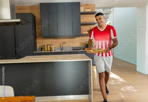 Konstfotografi African American man wearing striped tee walking in kitchen holding board with t