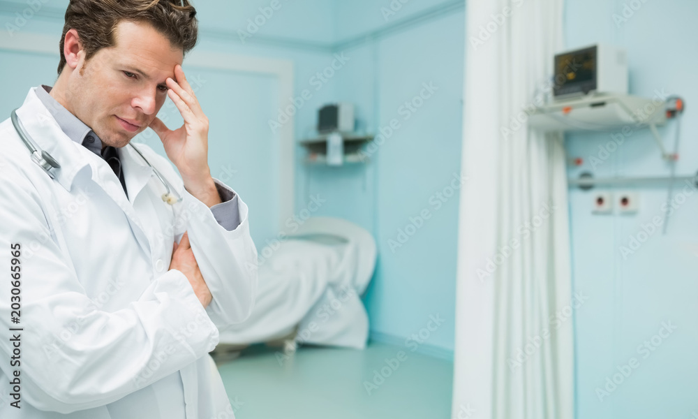 Fototapeta premium Male doctor wearing white coat, stethoscope, standing left, holding temple, viewing bed, copy space