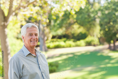 Senior man standing on left in park, wearing light button-up shirt, trees and manicured lawn