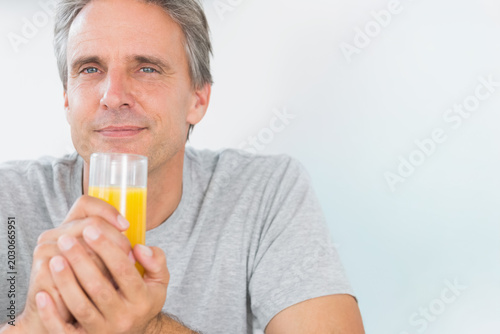 Middle-aged man sitting against pale wall holding clear glass of orange juice, wearing gray t-shirt