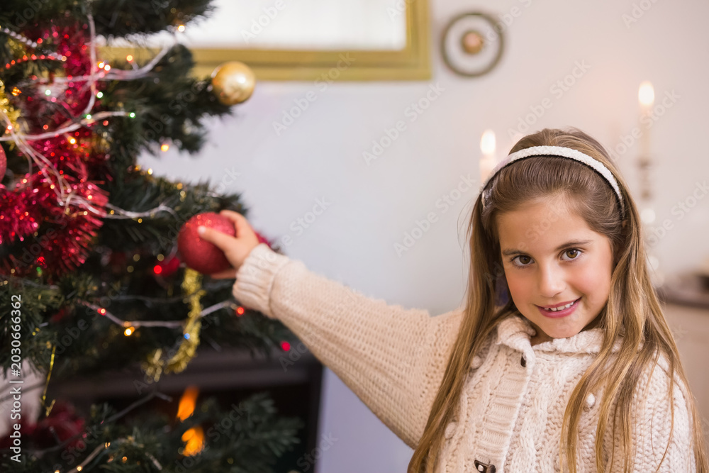 Fototapeta premium Placing large red bauble on Christmas tree in living room, child girl wearing knit sweater headband