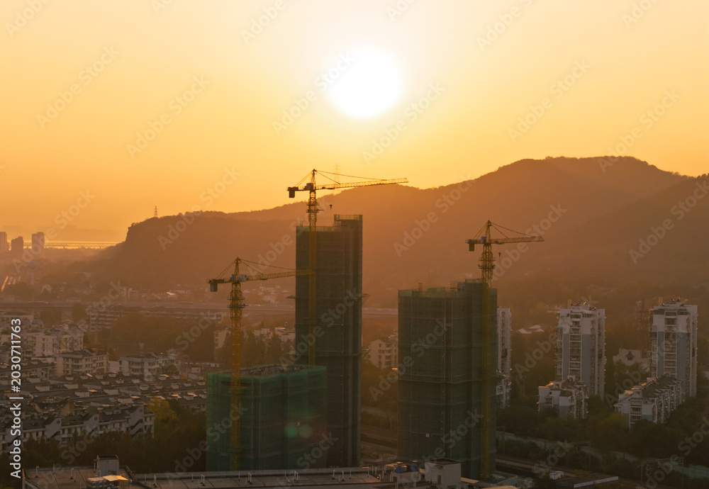 Fototapeta premium Building cranes silhouette against a vibrant golden sunset sky over a developing city nestled among distant mountains