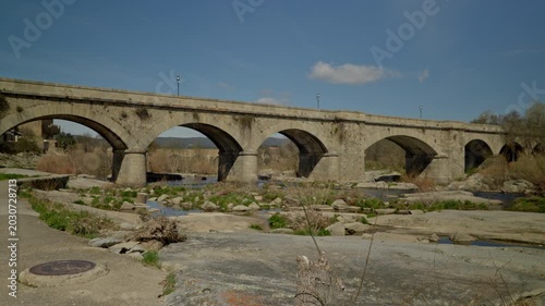 Ancient stone arch bridge crossing a low water river with rocks and dry veget...