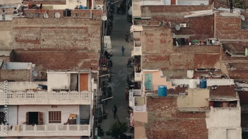 Aerial view of a narrow street in a densely populated Indian city, with people walking and motorcycles parked along the old, weathered brick buildings and rooftops in the warm afternoon light