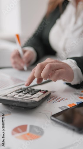 Closeup of female hands calculating data and reviewing business analytics on printed charts. Office workspace with calculator, smartphone, and financial documents for budget analysis and planning