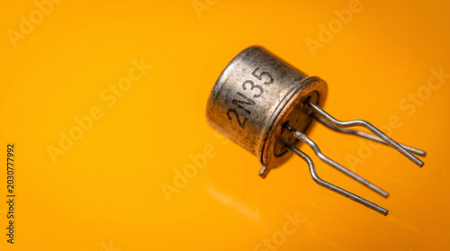 Single vintage germanium transistor with three metal leads splayed from metallic cap on amber background, semiconductor revolution and computing history concept.