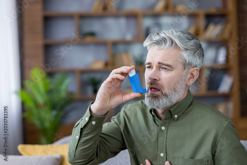 Senior man with gray beard using an inhaler at home to relieve asthma or respiratory distress, showing shortness of breath, discomfort, and self-care treatment