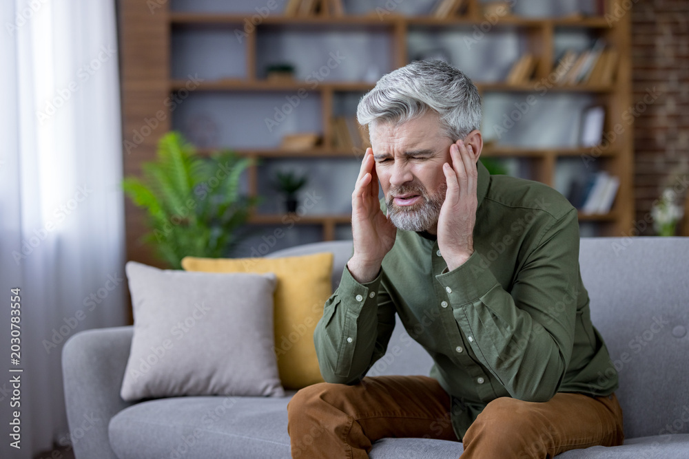 Fototapeta premium Mature man sitting on a sofa at home, experiencing intense headache or migraine pain, holding hands to temples with a pained expression, symbolizing stress, health issues, and discomfort