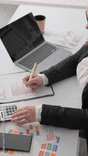 Focused female accountant calculating numbers on printed charts, graphs, surrounded by laptop, smartphone. Office workspace setup for financial planning, business analytics, data driven decision