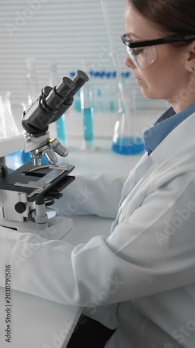 A woman scientist examines samples under a microscope in a bright laboratory environment, with test tubes and beakers in the background. Scientific research and discovery in progress