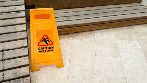 Yellow caution sign indicating wet floor placed on tiled surface near wooden steps, warning of potential slipping hazard in indoor environment
