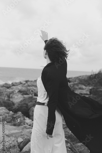 Foto Wind swept woman on a rocky coast in a flowing white dress and dark coat raises arms toward the sea, a bold windblown moment of freedom