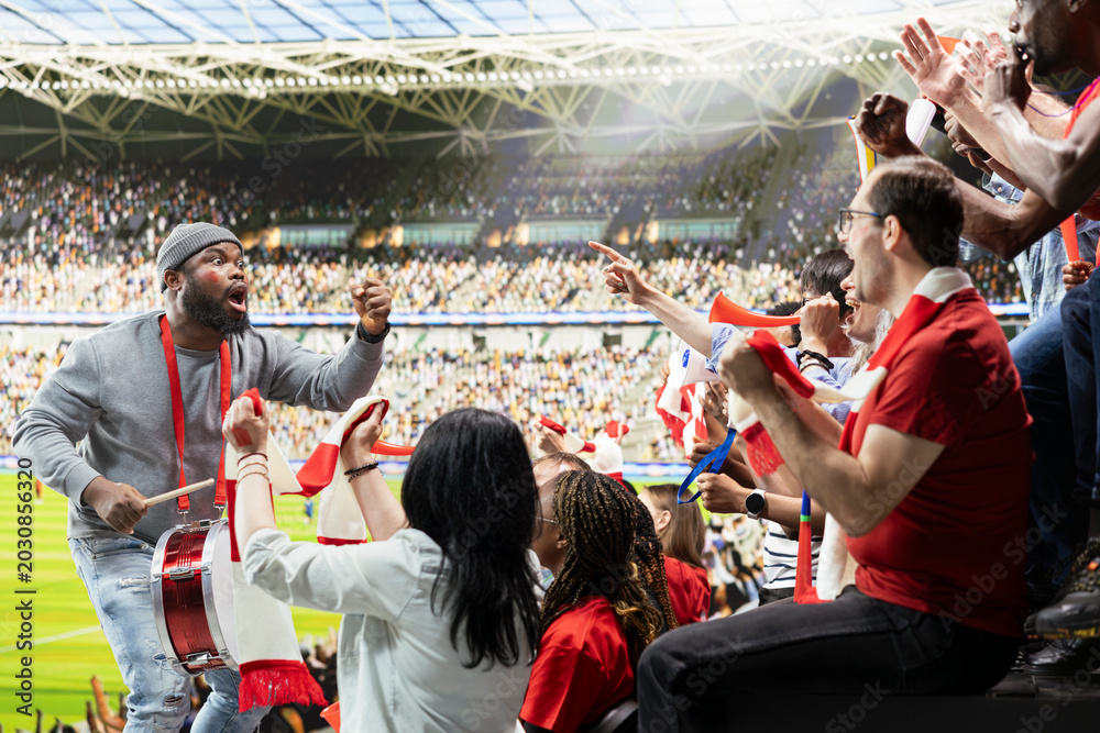 Obraz premium Supporter group leader creating intense energy by playing fan drums and leading chants from fan section during football match. Diverse sports fans with face paint and raised scarves in passion.