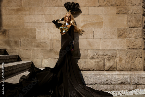 Obraz na plátně Majestic female model in avant-garde flowing black gown with metallic gold armor and elaborate feather headdress posing against a textured stone wall in a high-fashion fantasy editorial