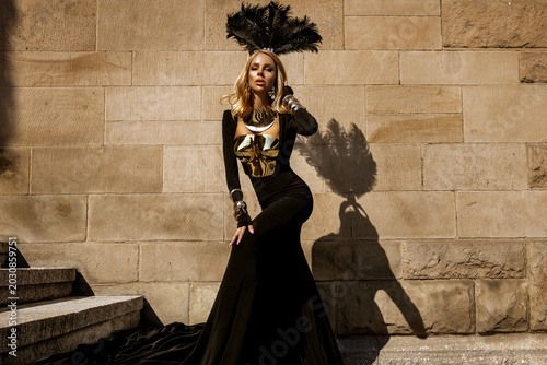 Fotografi Majestic female model in avant-garde flowing black gown with metallic gold armor and elaborate feather headdress posing against a textured stone wall in a high-fashion fantasy editorial